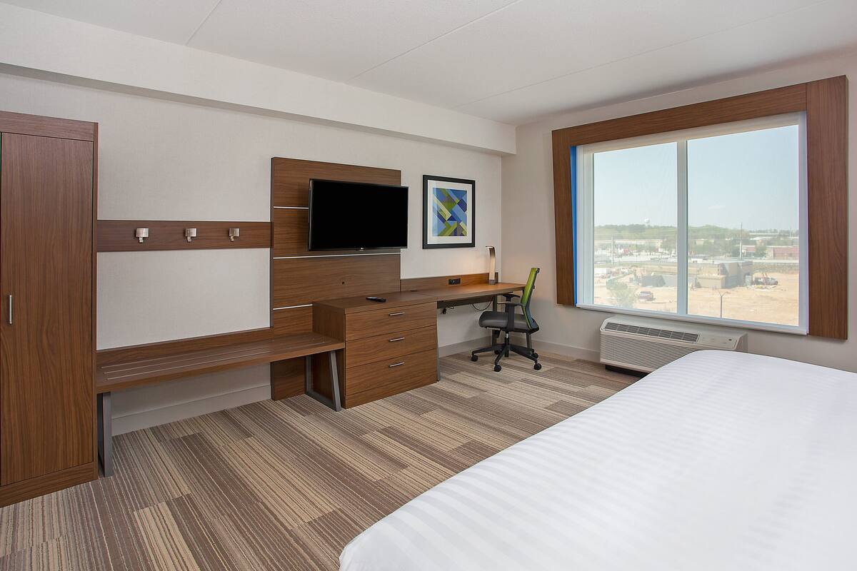 A hotel room with a bed, a desk with a flat-screen TV, a chair, and a large window.