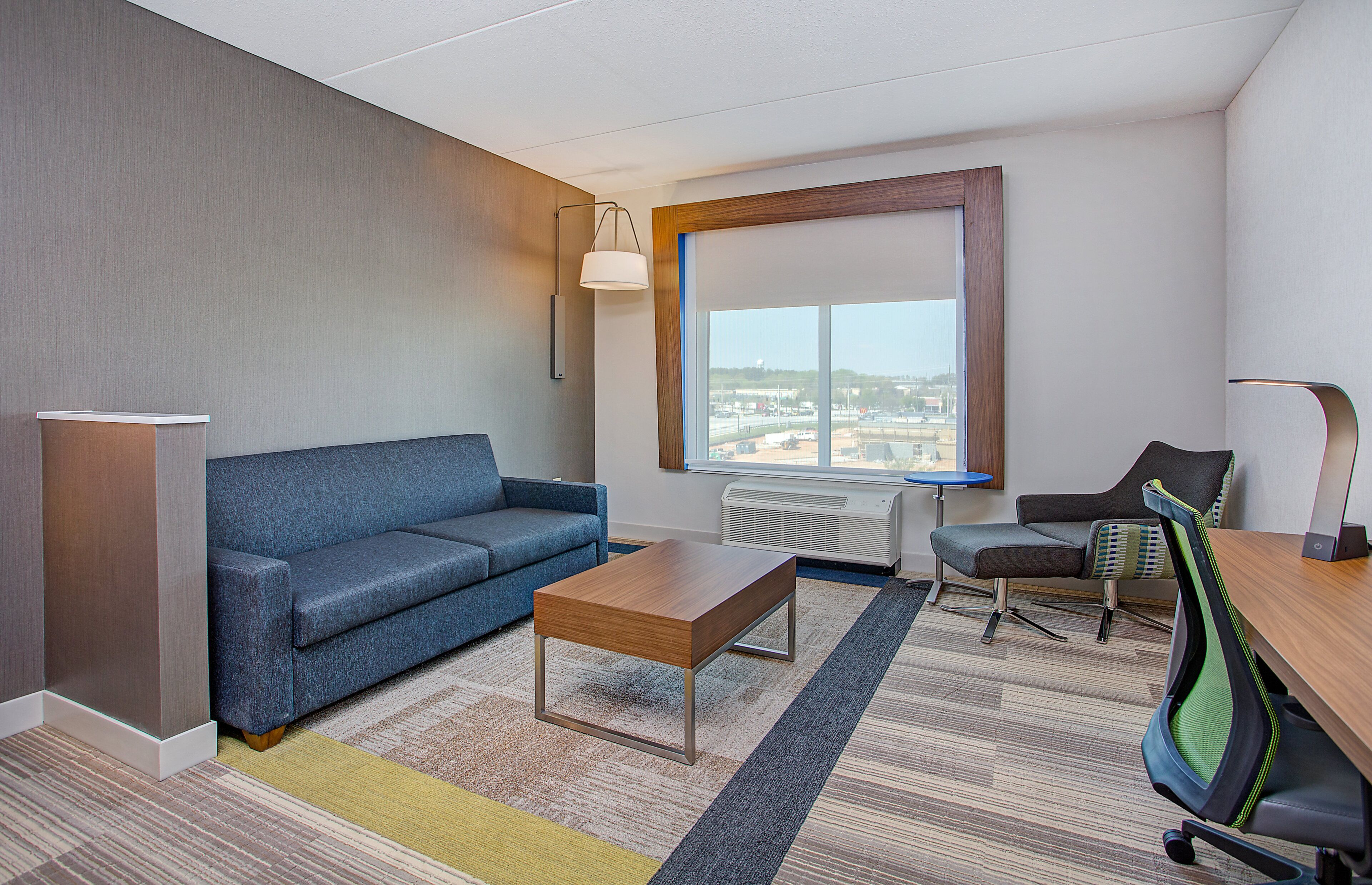 Een moderne hotelkamer met een blauwe bank, een houten salontafel, een bureau met stoel, een groot raam met uitzicht op een stadsbeeld, en een staande lamp.