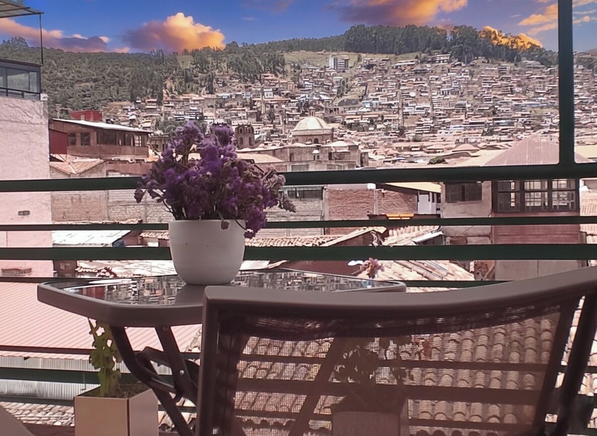 Un balcón con una mesa y sillas, una planta en maceta y vistas de una ciudad densamente poblada con edificios y una colina al fondo.