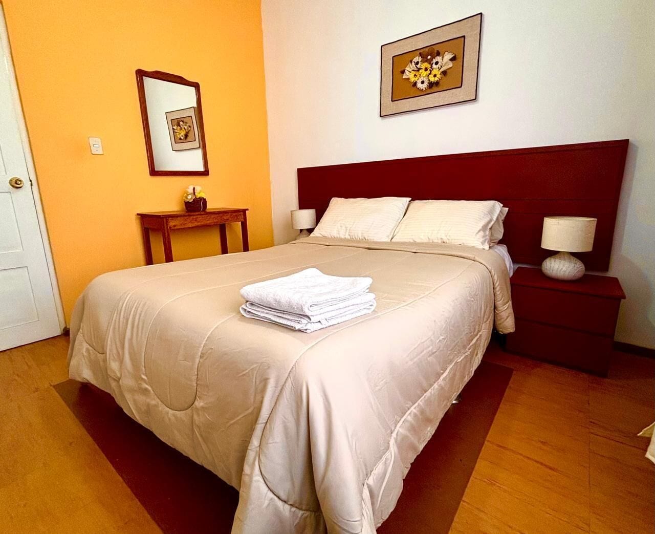 A neatly made bed with a beige bedspread and pillows, a small bedside table with a lamp, and a framed picture on the wall.