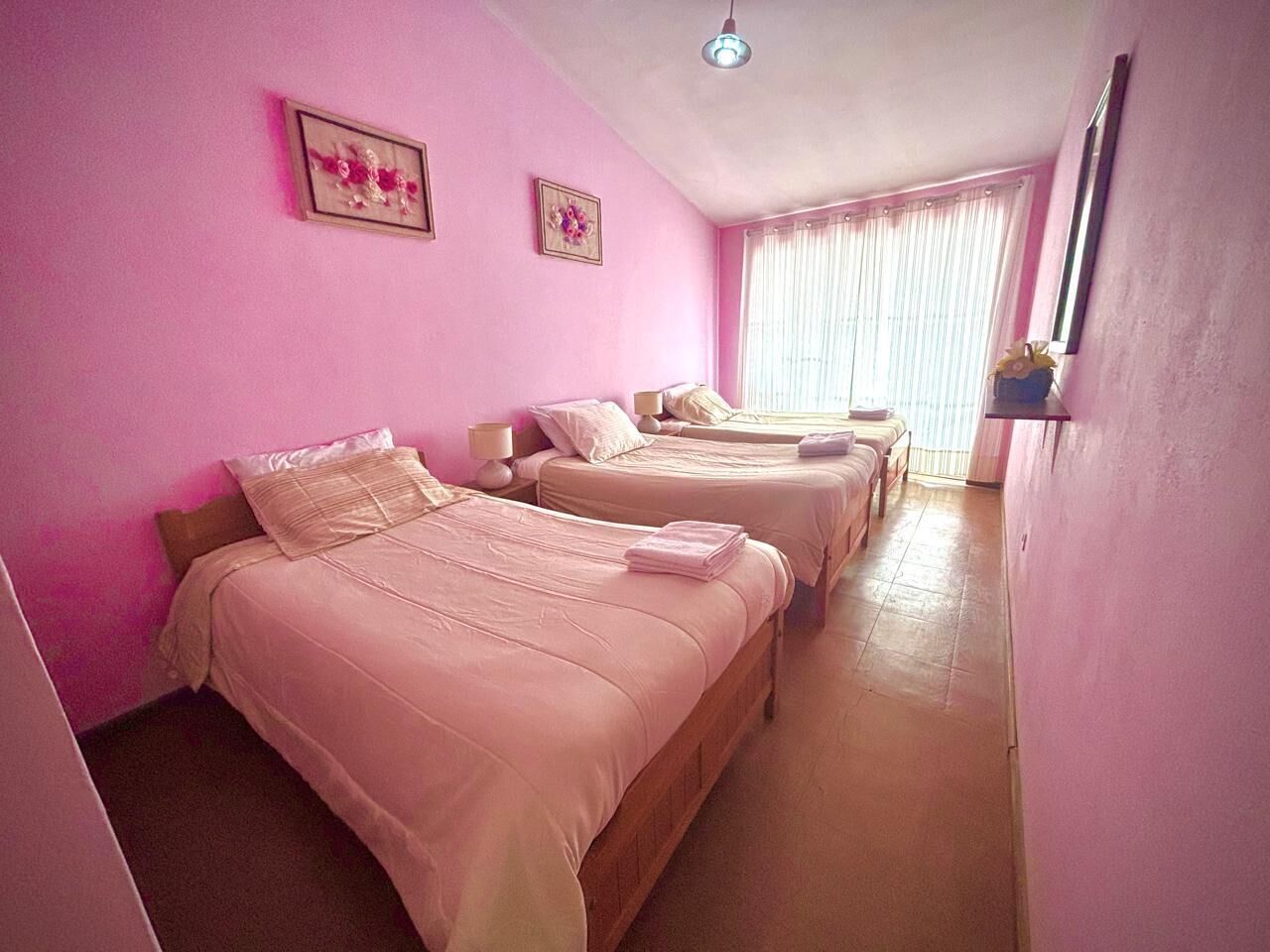 A room with pink walls, two beds with matching linens, a bedside table with a lamp, and a window with curtains.