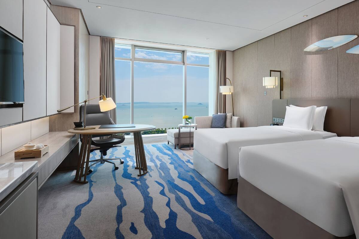 A hotel room with a large window, a desk with a lamp, two beds, and a view of the sea.