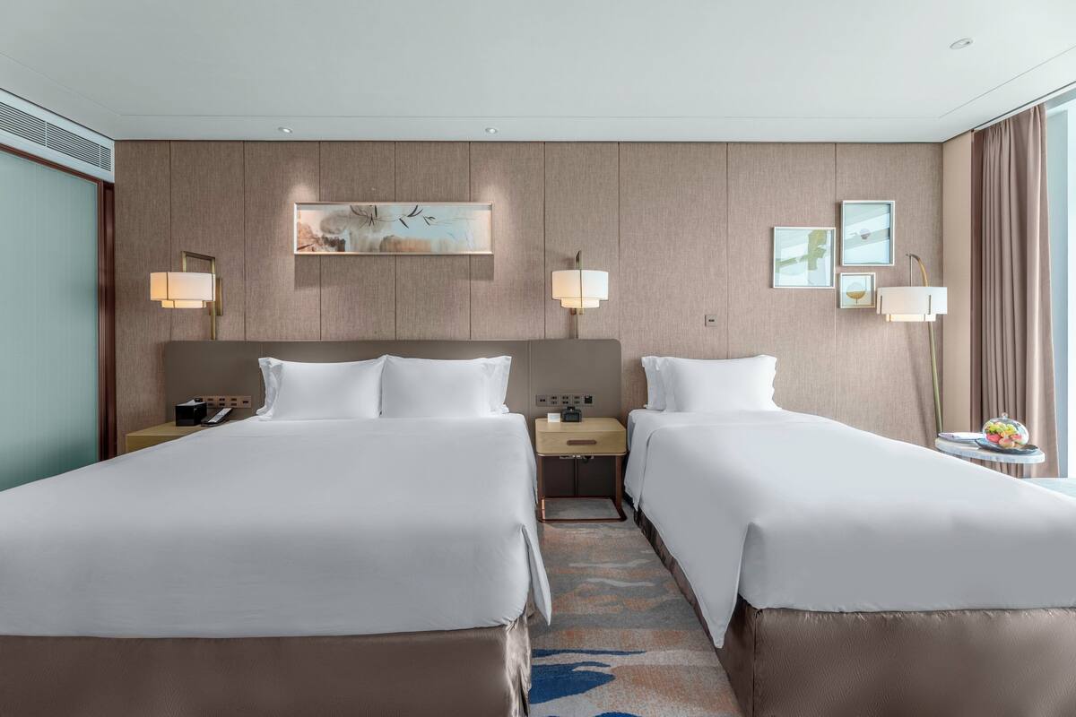 A hotel room with two beds, a nightstand with a lamp, a painting on the wall, and a window with curtains.