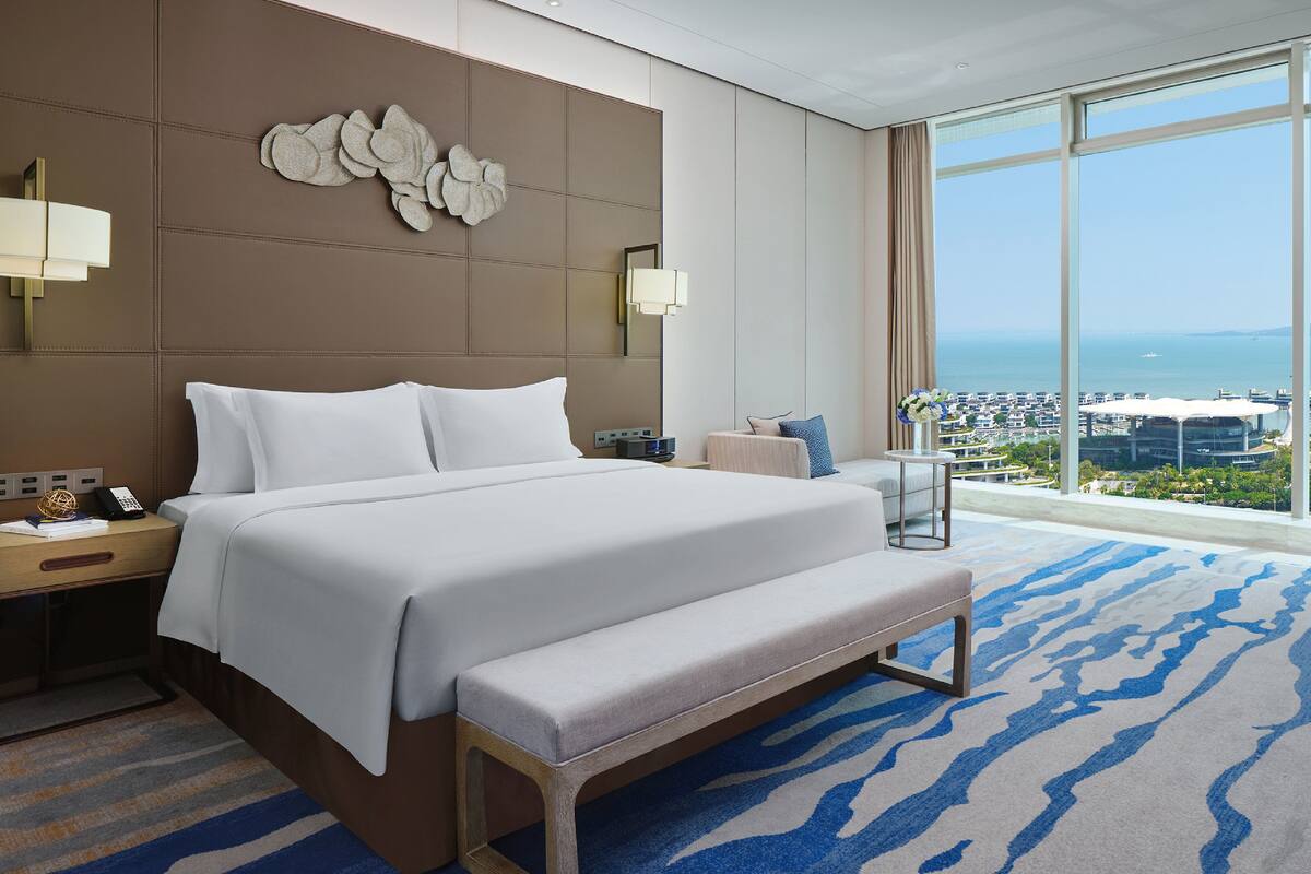 A hotel room with a large bed, a bench, a desk, and a view of the city and sea.