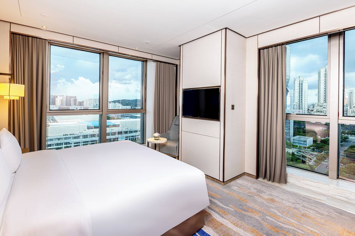 A modern hotel room with a large bed, a flat-screen TV, a small table, and a city view through large windows.