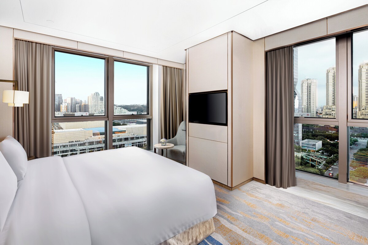 Hotelkamer met een groot bed, een flatscreen televisie, een klein tafeltje en uitzicht op de stad door grote ramen.