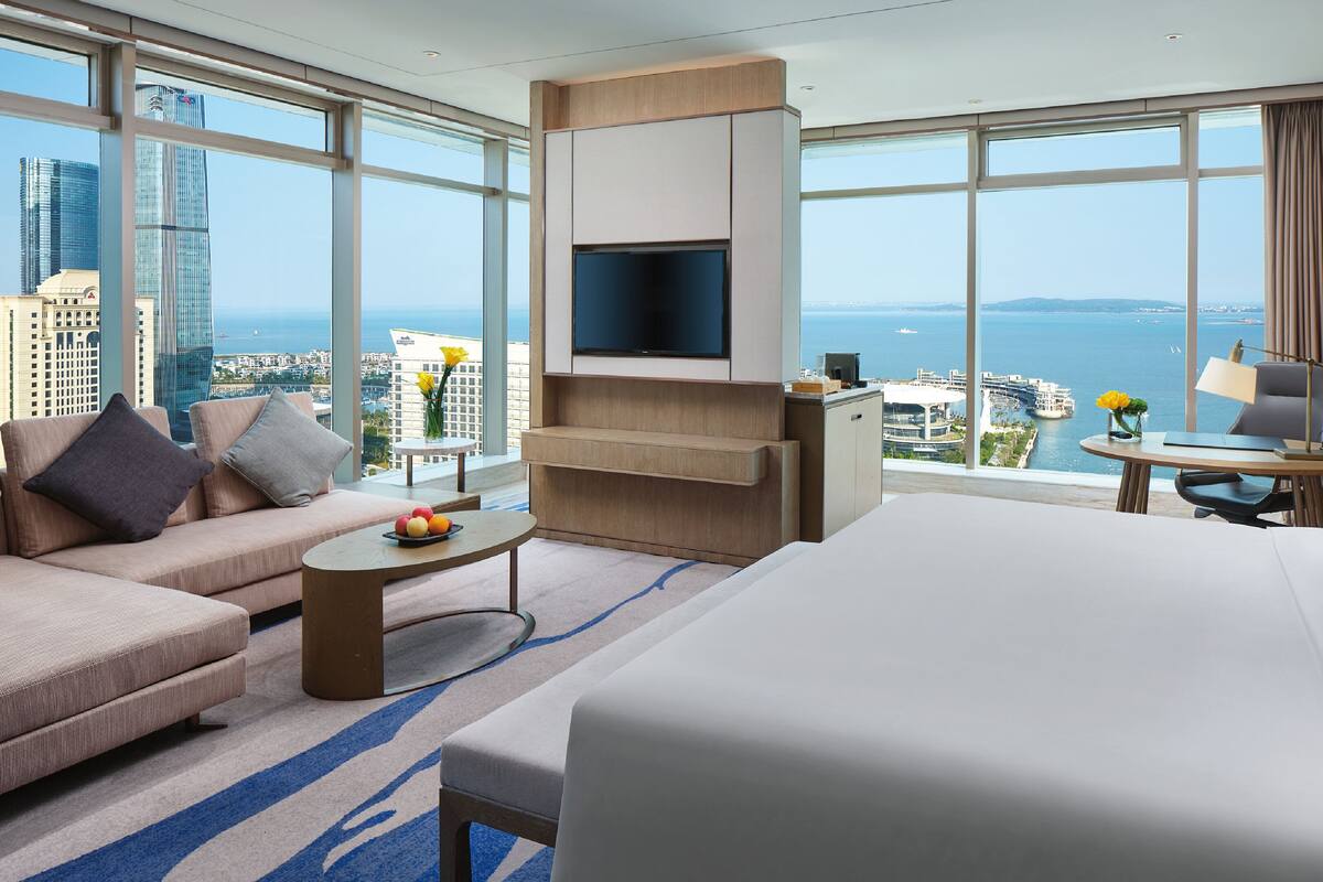 A modern hotel room with a large bed, a sofa, a TV, and a desk with a view of the city and sea.