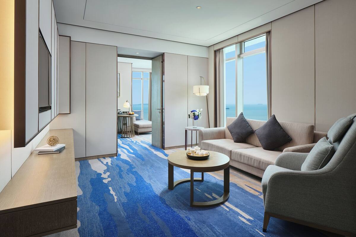 A modern hotel room with a sofa, a round coffee table, a desk, and a view of the sea.