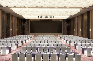 Un amplio salón de conferencias con filas de sillas blancas, mesas con botellas de agua y una pantalla grande al frente.