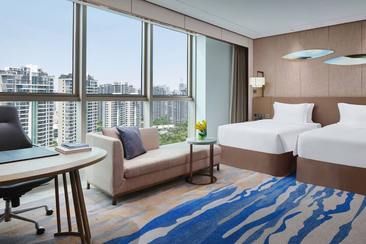 A hotel room with two beds, a desk, a chair, and a view of the city.