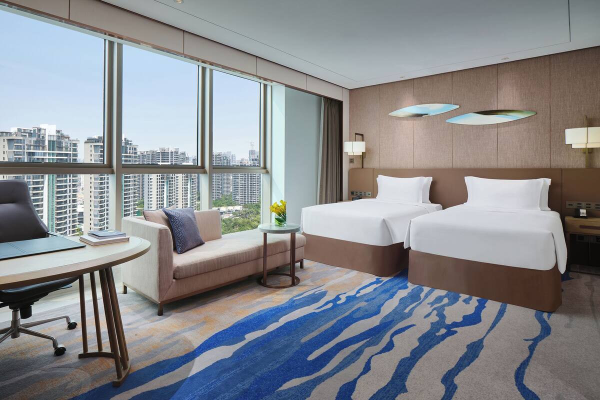 A hotel room with a large window, two beds, a desk, and a sofa.