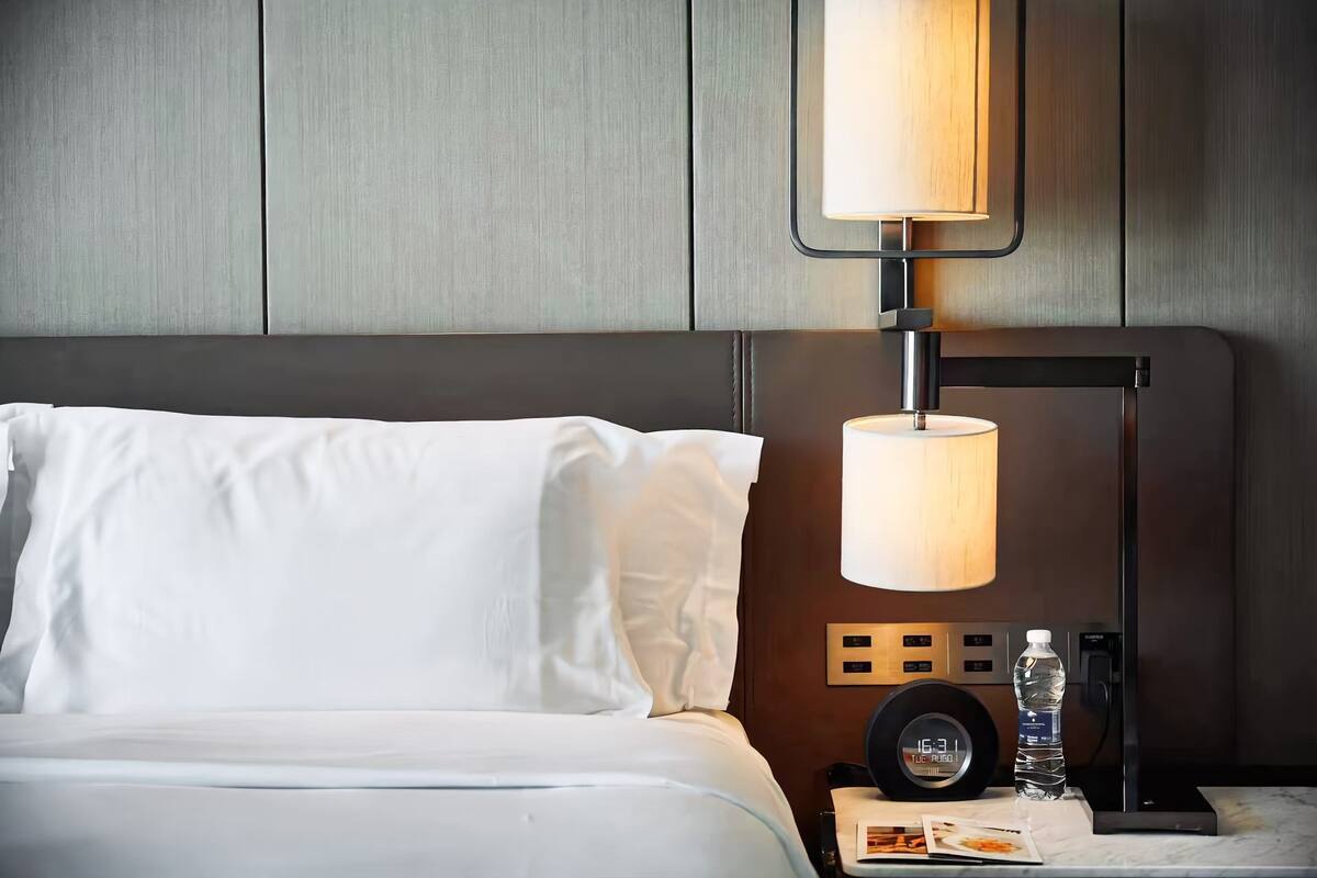A neatly made bed with white linens, a wall clock, a water bottle, and a lamp on a bedside table.