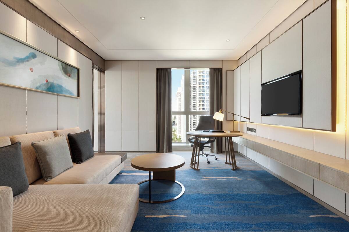 A modern hotel room with a sofa, a round table, a desk, a TV, and a large window with a city view.