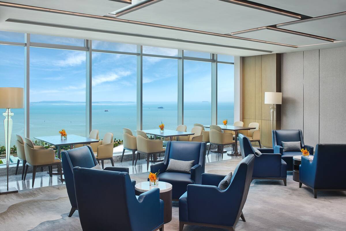 A modern hotel lobby with large windows offering a view of the sea. There are several seating areas with tables and chairs.