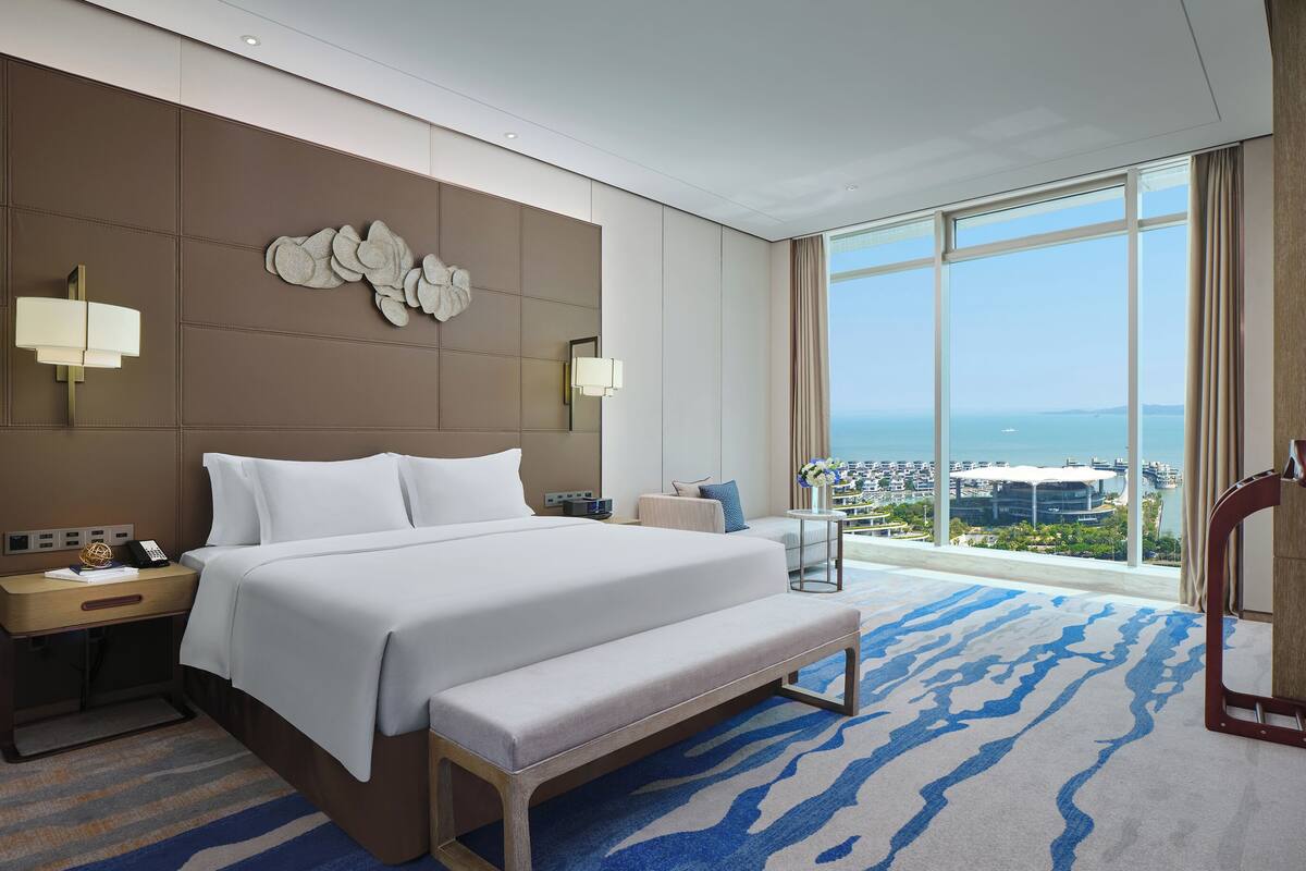 A hotel room with a large bed, bedside tables, a bench, and a view of the city and sea.