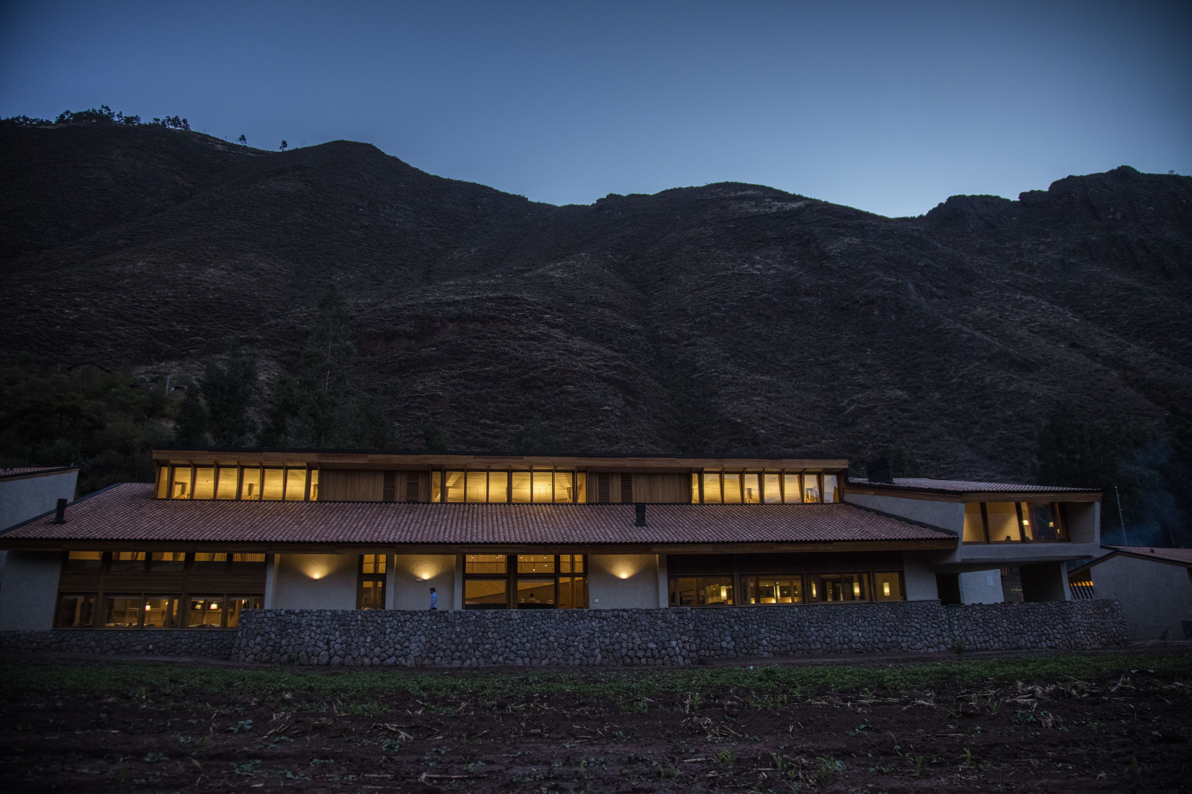 A modern two-story house with large windows and a stone foundation, set against a mountainous backdrop.