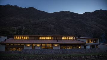 A modern two-story house with large windows and a stone foundation, set against a mountainous backdrop.