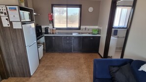 A compact kitchen with a microwave, refrigerator, and cabinets. There is a window above the sink, and a blue sofa is visible in the foreground. - Windana Cottages (Hawker)