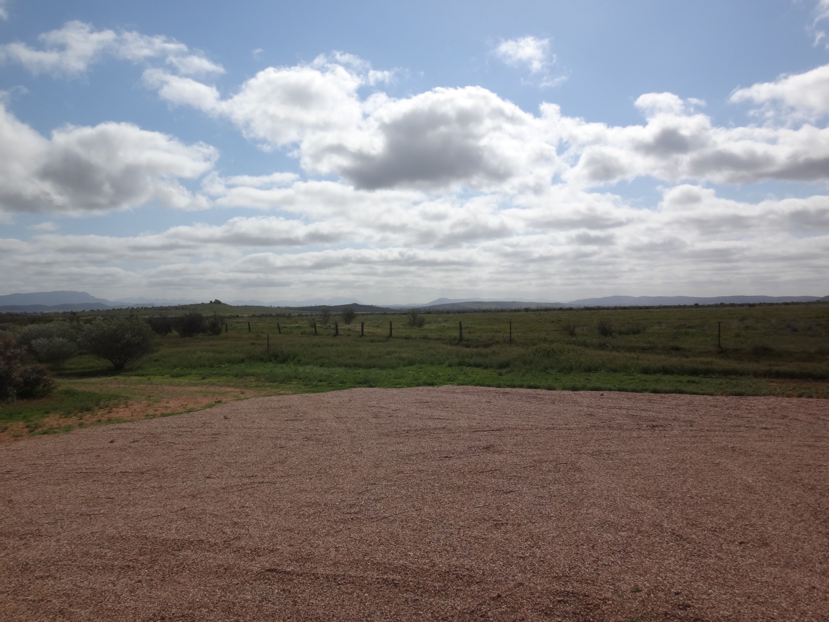 A rural landscape with a gravel path leading into a field, surrounded by low vegetation and distant hills under a partly cloudy sky.