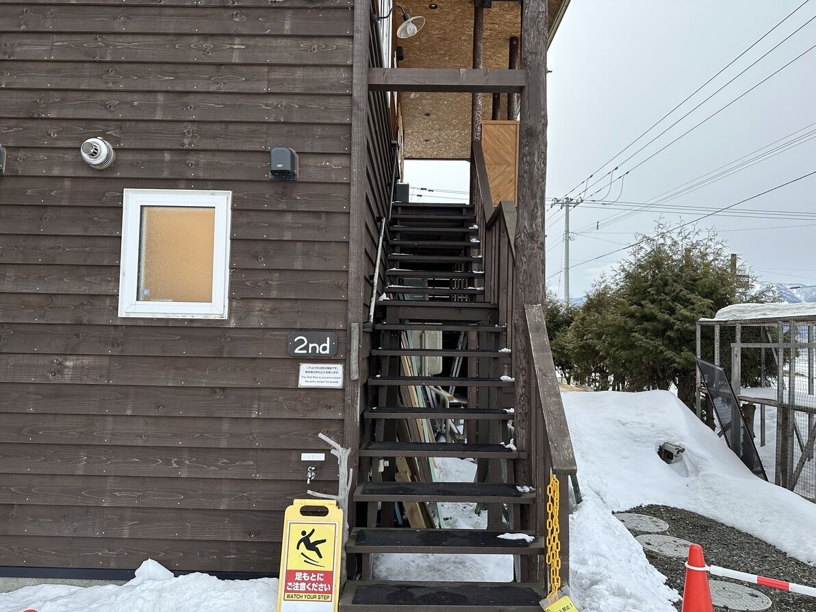 A wooden building with a staircase leading to a second-floor entrance, a fire hose reel, and a yellow caution sign.