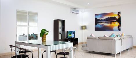 A modern living room with a glass dining table, black chairs, a grey sofa, and a large painting of a sunset over a lake.