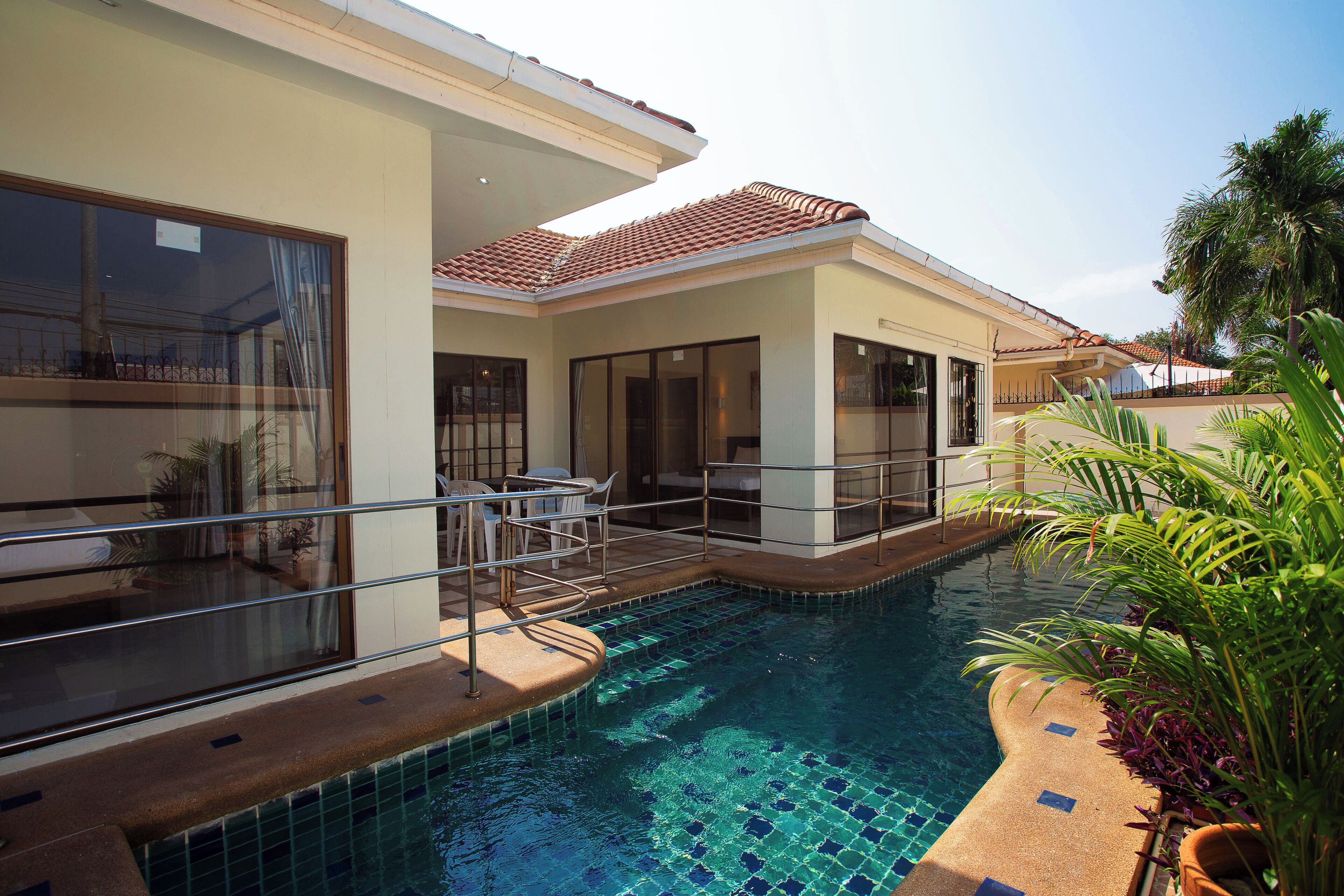 A swimming pool with a tiled edge, a covered area with chairs, and a building with large windows and doors.