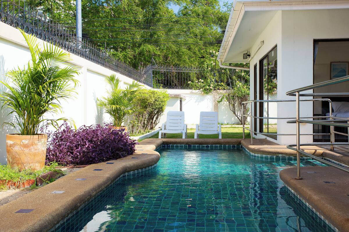 A swimming pool with a tiled edge, surrounded by a white wall and a metal fence. There are two white lounge chairs and a potted plant near the pool.