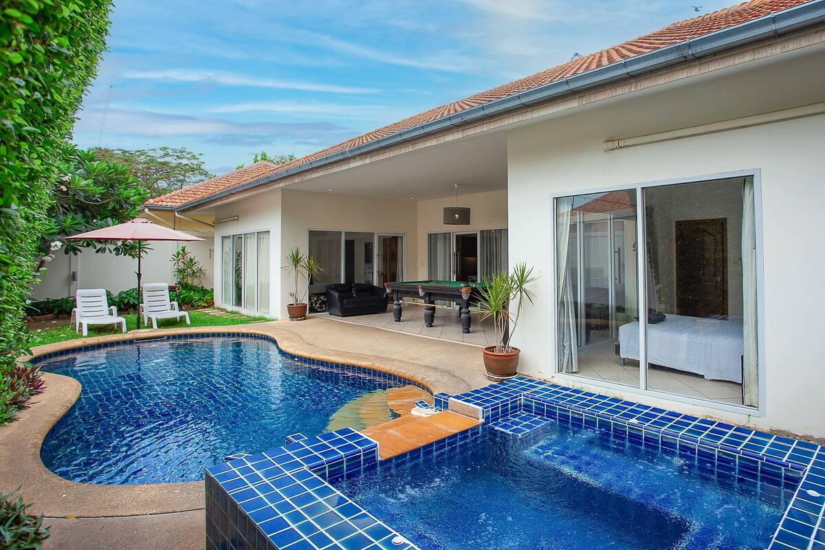 A swimming pool with blue tiles, a covered patio with chairs, and a bed in a room with a sliding glass door.