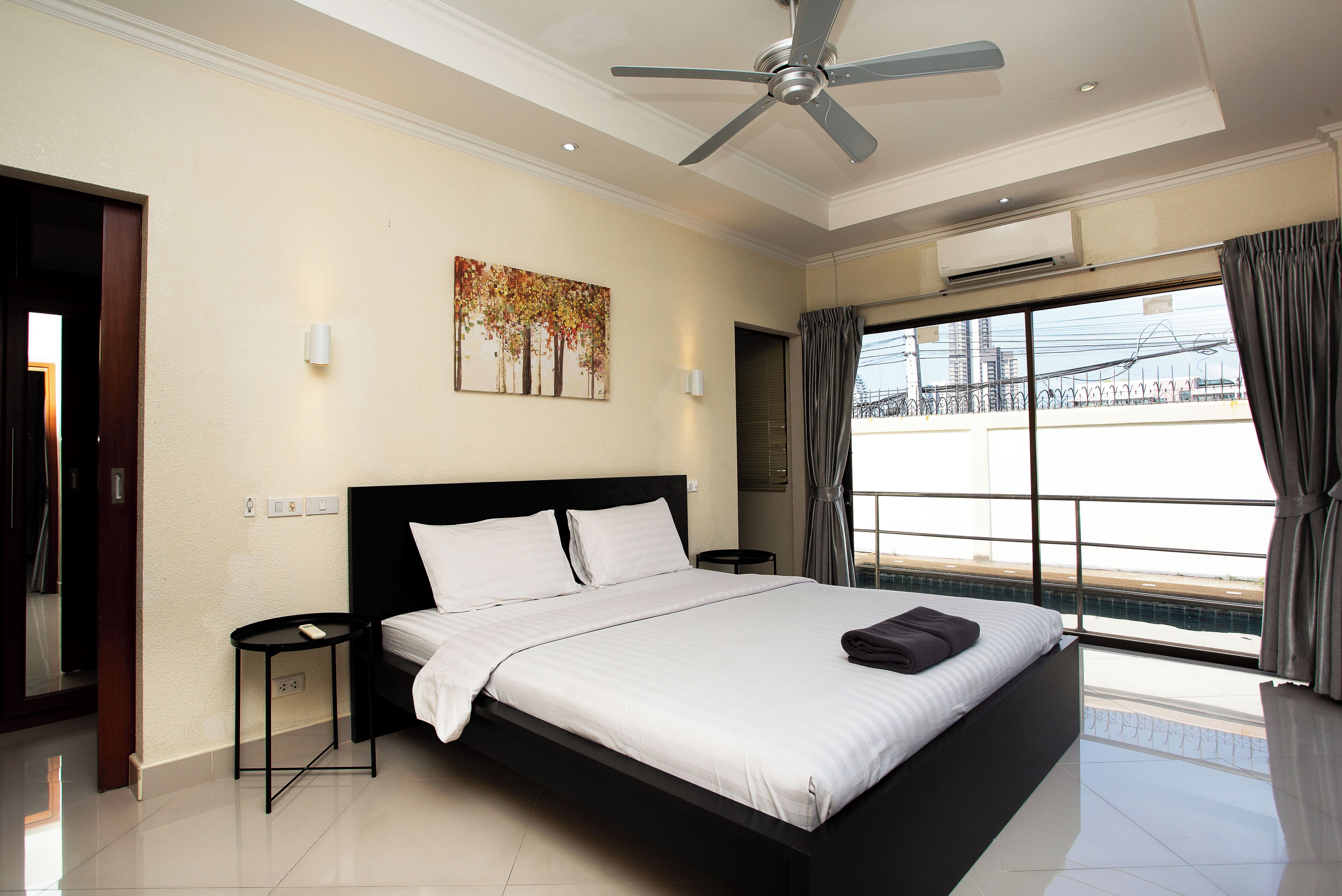 A modern hotel room with a large bed, a ceiling fan, a painting on the wall, and a view of the city through the window.
