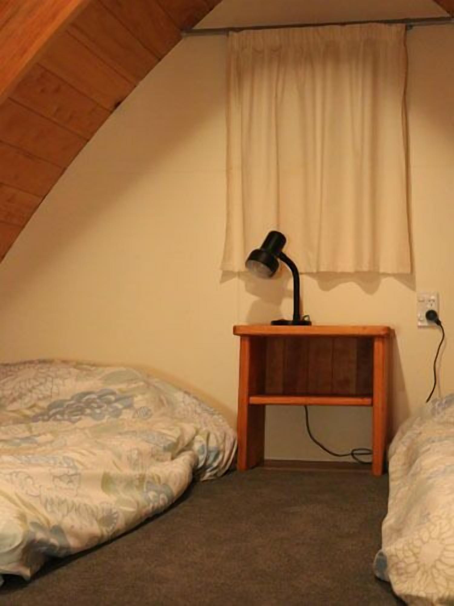 Une petite chambre agréable, dotée de deux lits simples, d’une table de chevet en bois avec une lampe et d’une fenêtre avec des rideaux.
