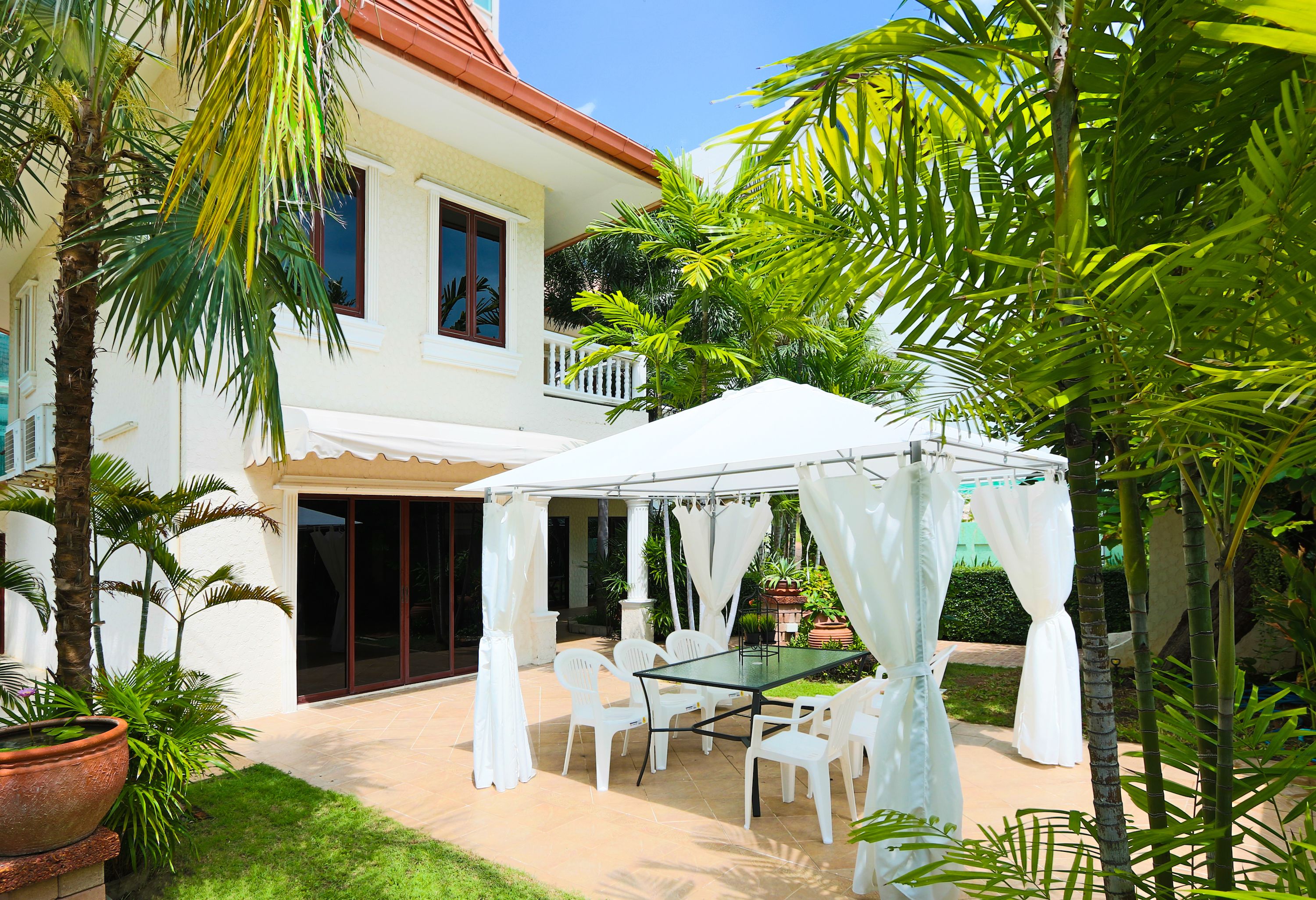 Une terrasse extérieure couverte, avec une table en verre et des chaises blanches, entourée d’une végétation luxuriante et d’un bâtiment blanc doté d’un toit rouge.