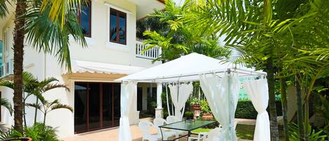Une terrasse extérieure couverte, avec une table en verre et des chaises blanches, entourée d’une végétation luxuriante et d’un bâtiment blanc doté d’un toit rouge.