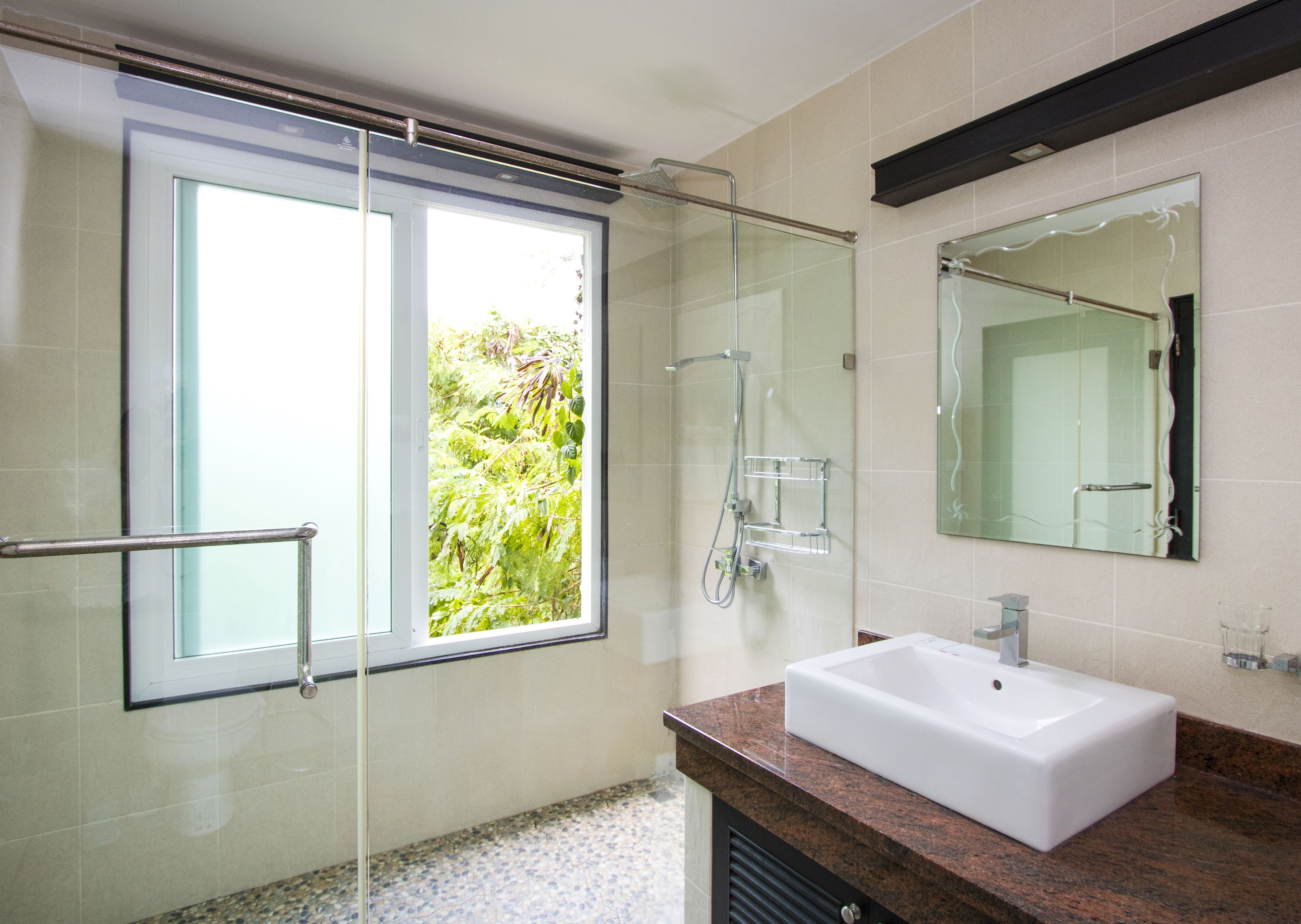 A modern bathroom with a glass shower enclosure, a large window with a view of greenery, a white sink, and a mirror above it.