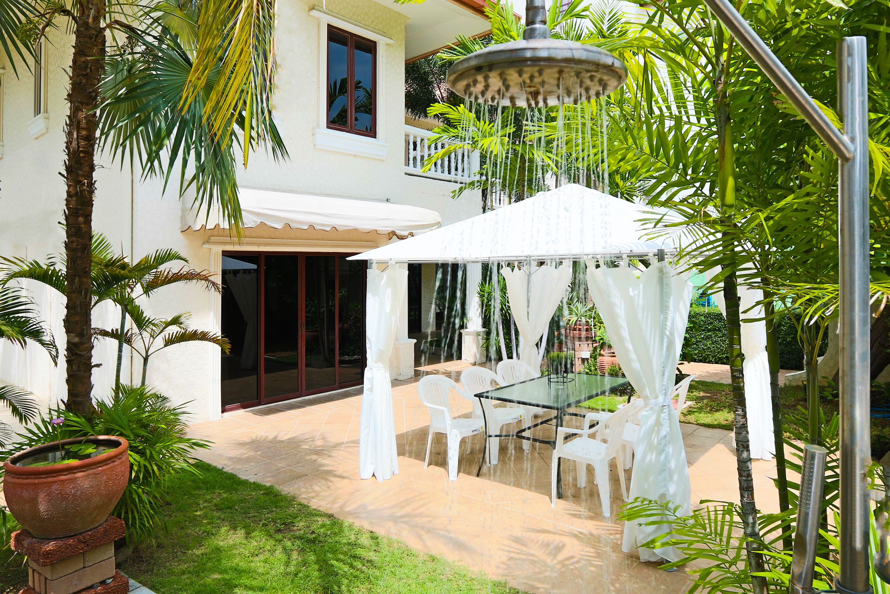 A covered outdoor seating area with a glass table and chairs, surrounded by palm trees and a white building.