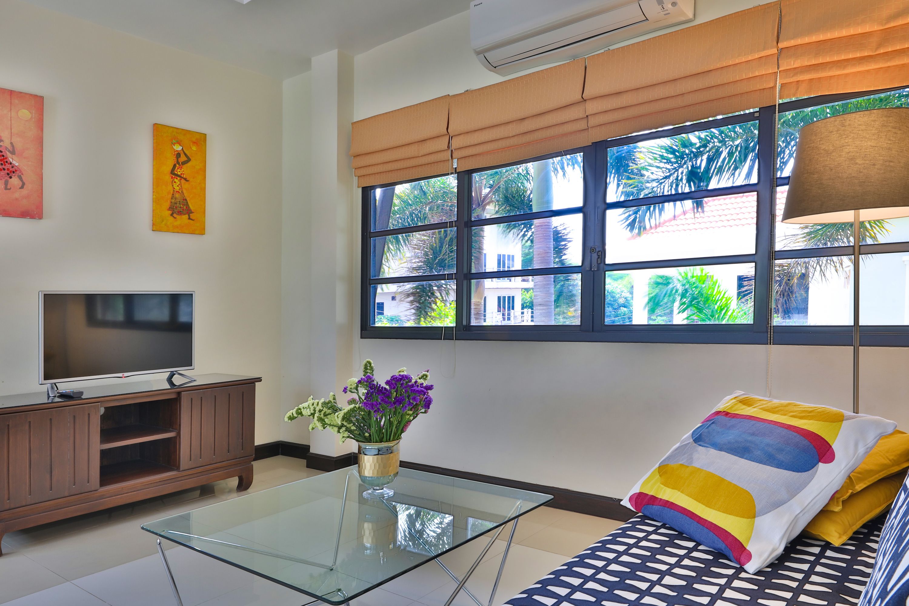 A living room with a glass coffee table, a TV on a wooden cabinet, a sofa with patterned cushions, and a window with blinds.