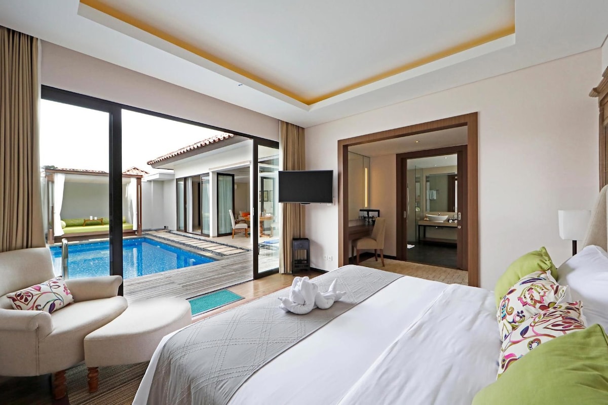 A hotel room with a bed, a chair, a TV, a bathroom, and a pool area.