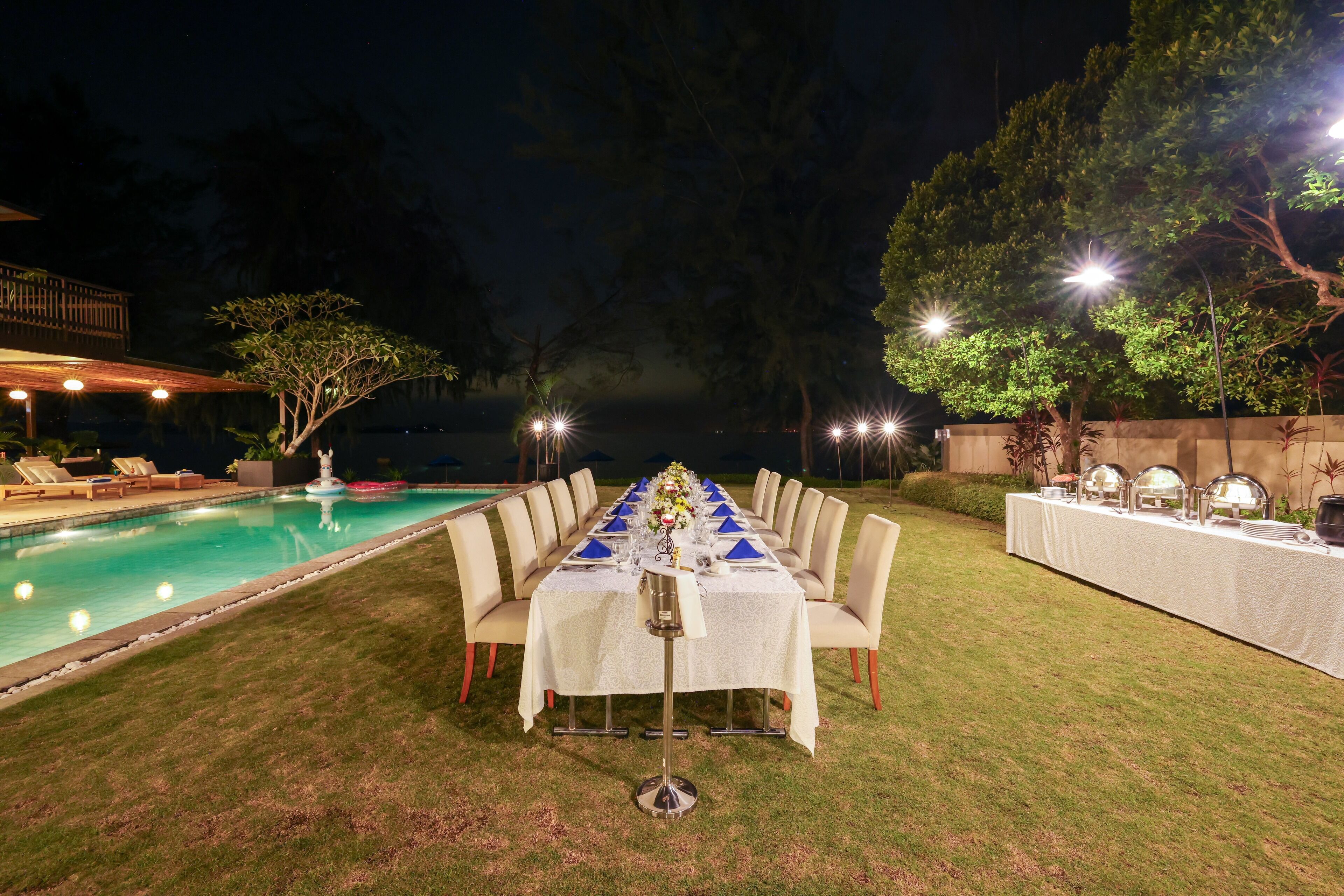 Un área de comedor al aire libre con piscina, mesas dispuestas para una comida y una iluminación cálida en la noche.