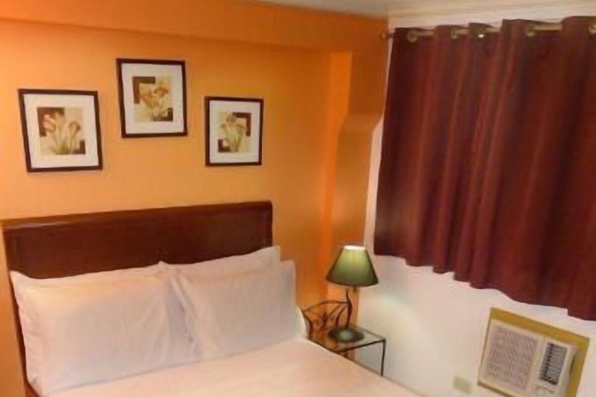 A neatly made bed with white linens and a red blanket, a bedside table with a lamp, and a wall-mounted air conditioning unit.