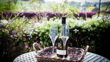 A wicker basket with a wine bottle and two glasses on a round table, set outdoors with a garden background.