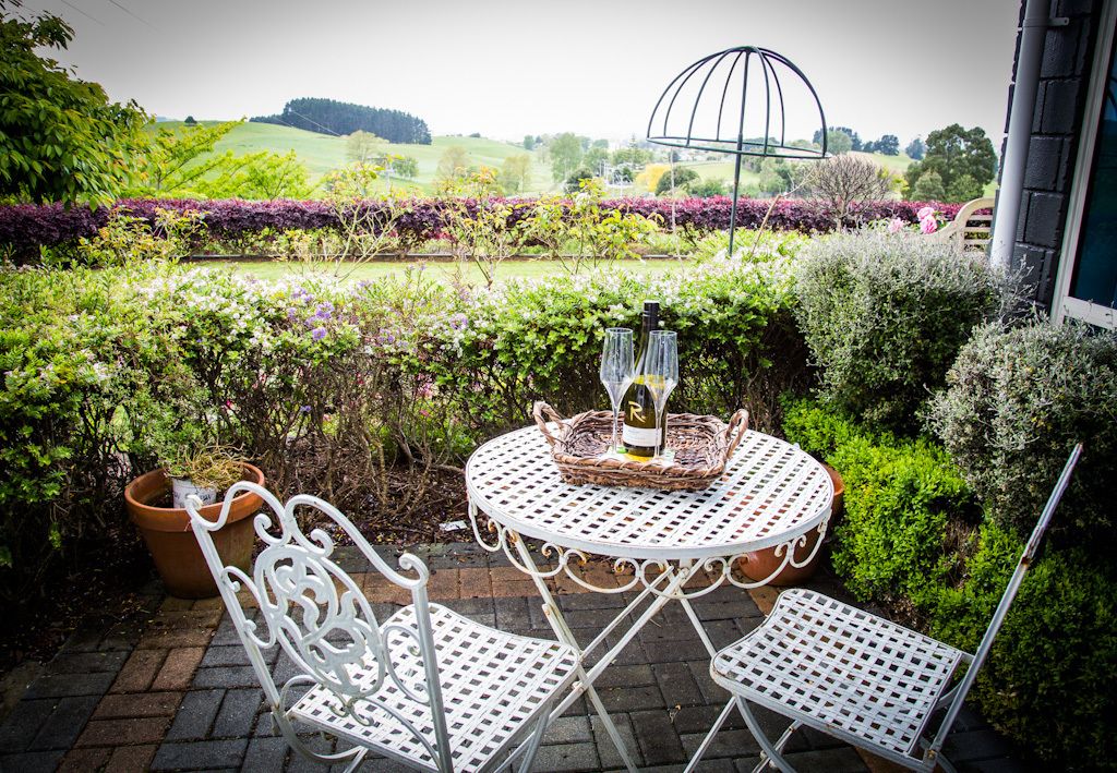 Une table ronde en métal, accompagnée de deux chaises blanches, une bouteille et un verre, est placée sur une terrasse en briques donnant sur un jardin.
