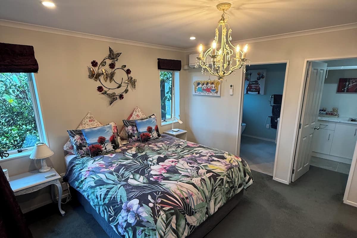 A bedroom with a floral bedspread, a chandelier, a desk with a lamp, and a view of the outdoors through a window.