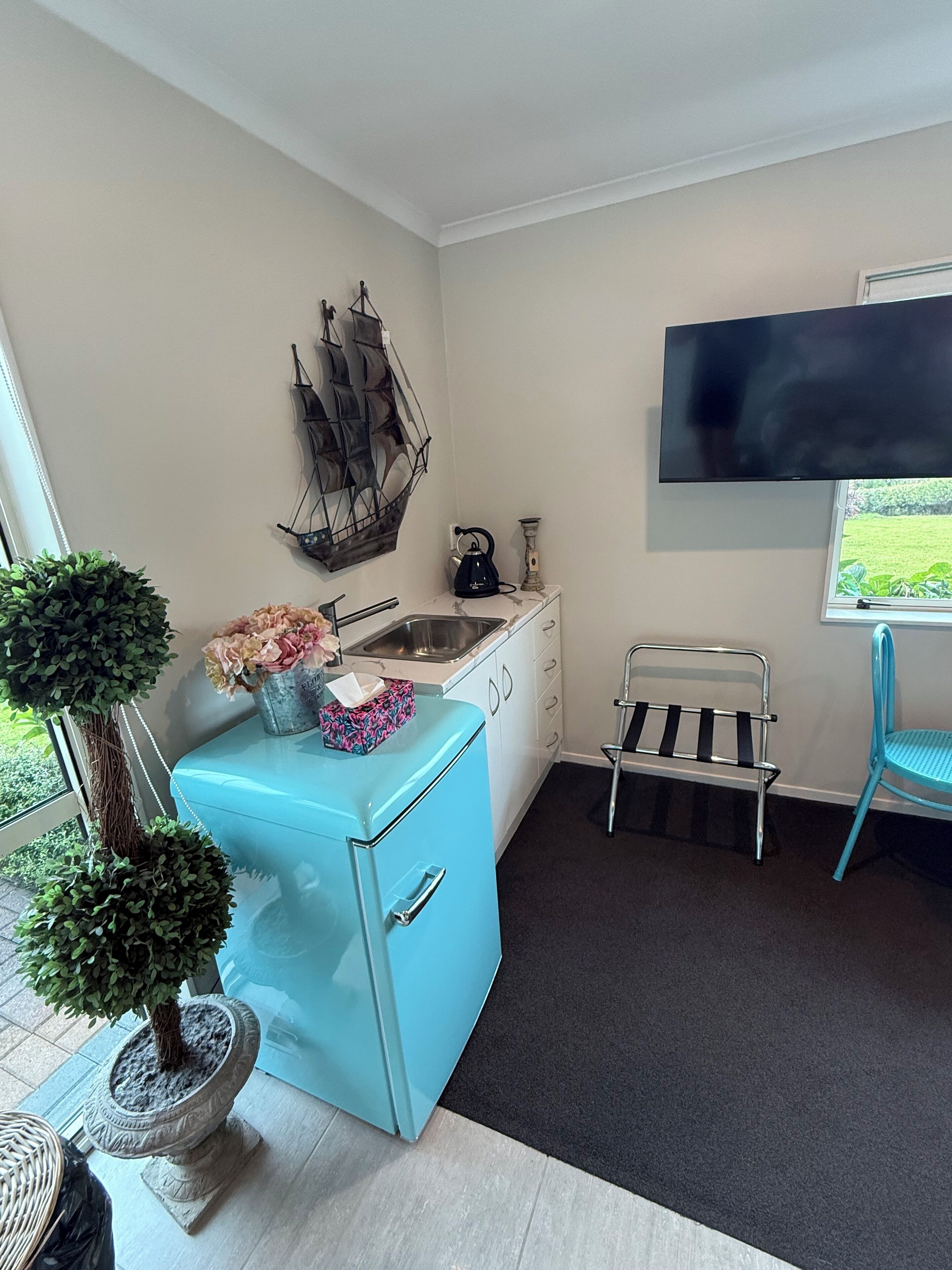 A compact kitchen with a blue refrigerator, a sink, and a kettle. There's a TV mounted on the wall, a window with a view, and a small bench.