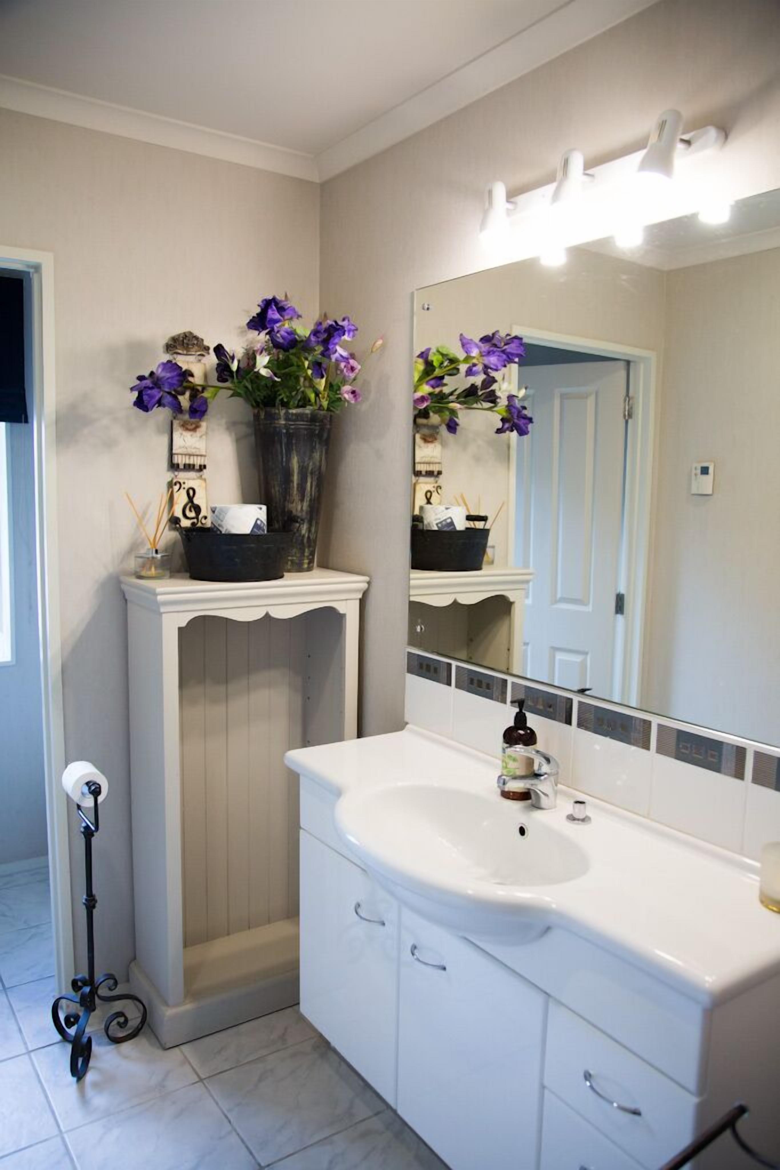 A bathroom with a white sink, vanity, and a mirror with lights. There is a vase with purple flowers on the vanity.