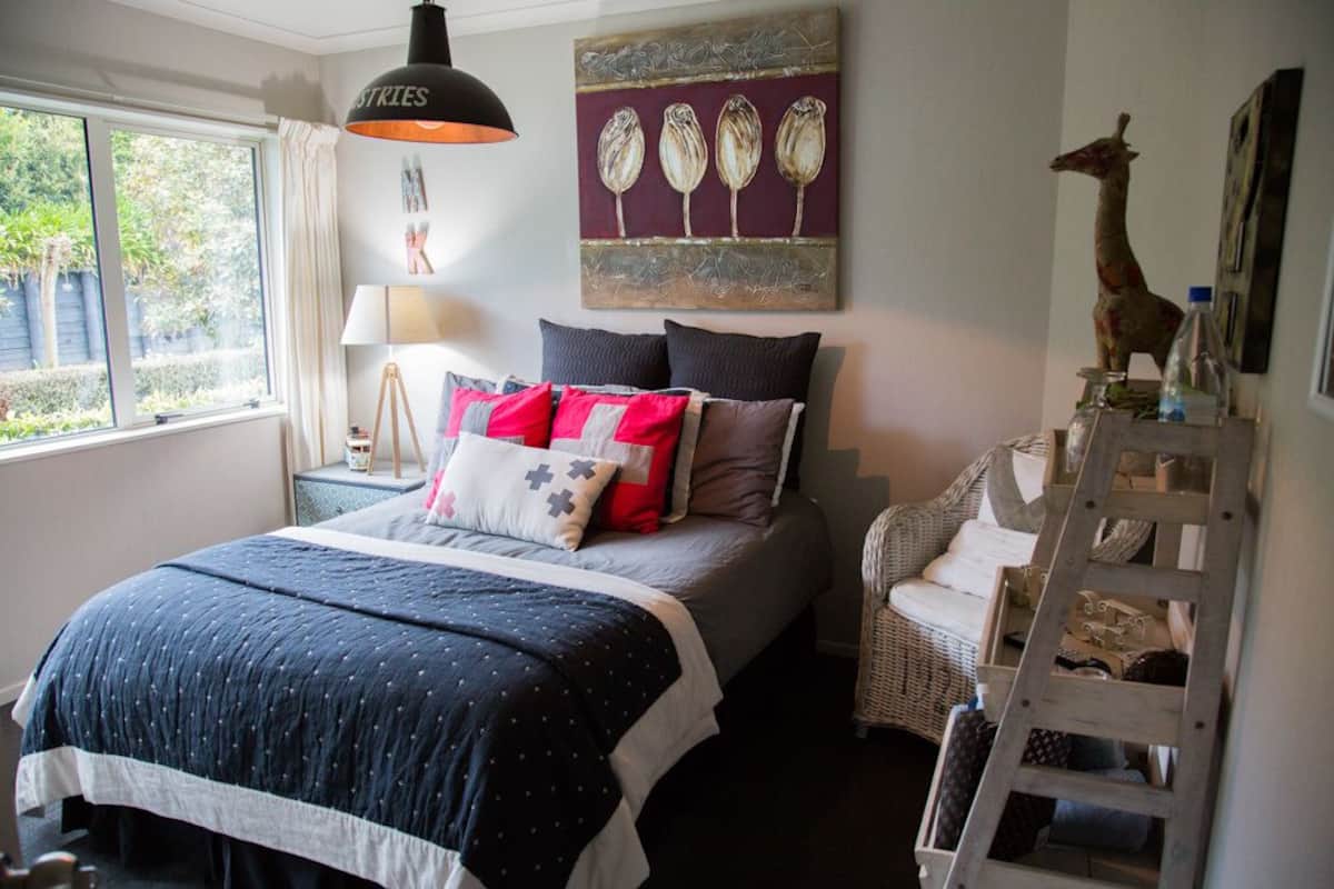 A bedroom with a bed, a nightstand, a lamp, a wicker chair, a ladder, and a wall with a painting.