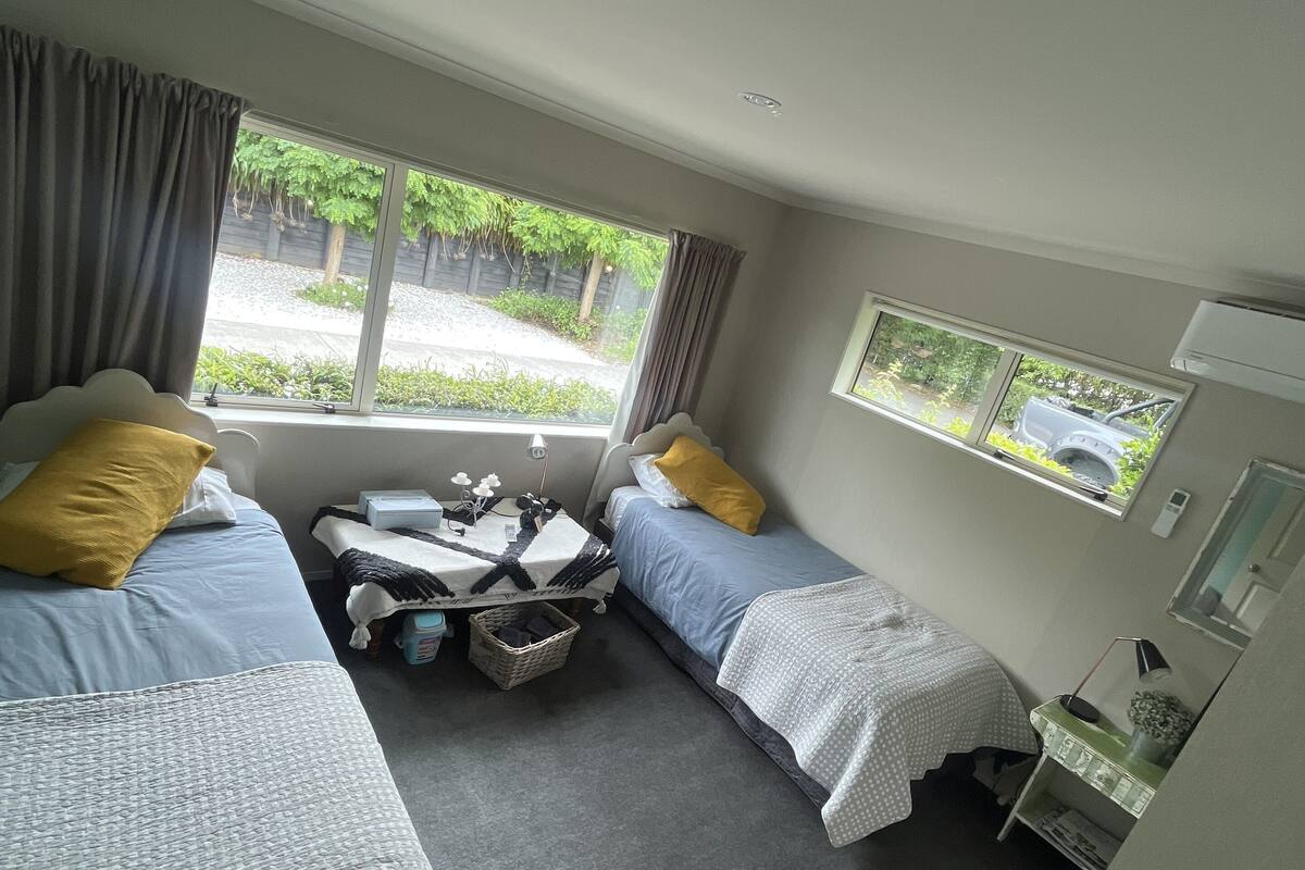 A small, well-lit room with two beds, a small table, and a window providing a view of the outdoors.