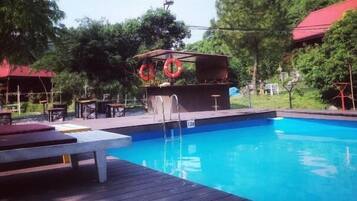 Relax by the pool surrounded by lush greenery and trees, with lounge chairs and a shaded seating area.