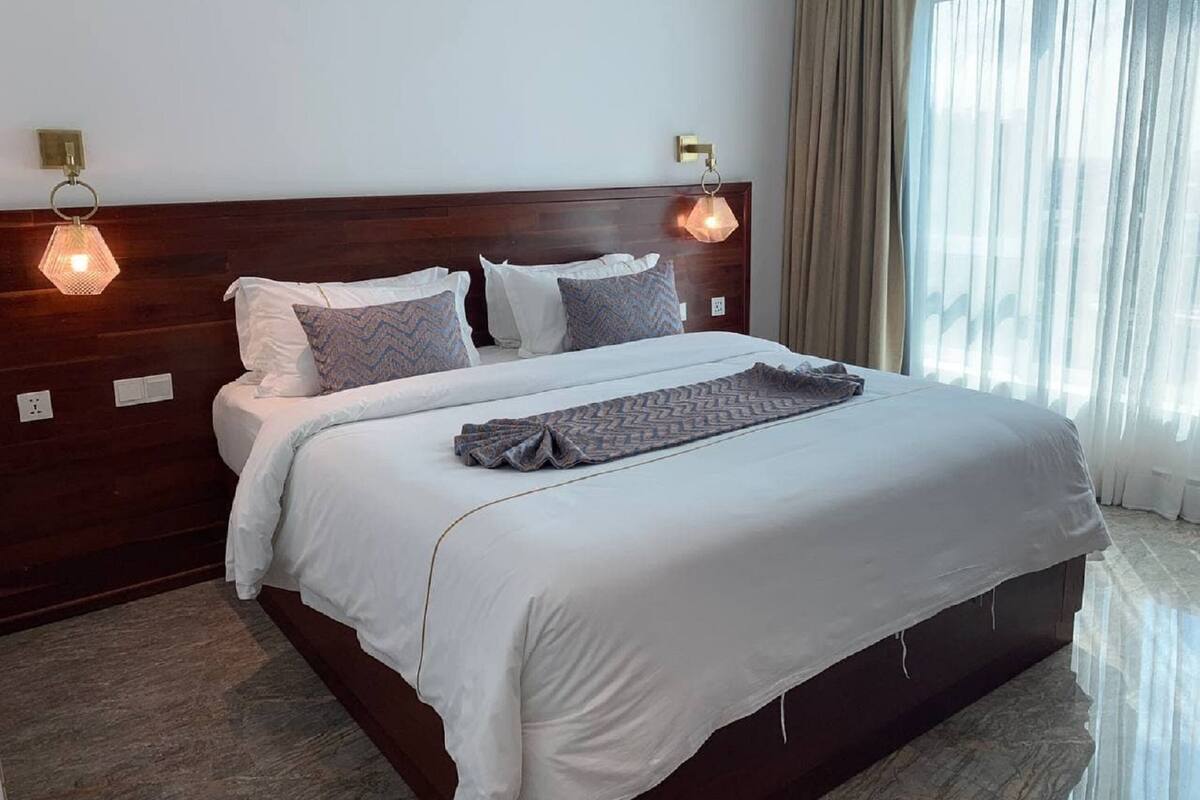 A hotel room with a large bed, two wall-mounted lamps, a wooden headboard, and a window with curtains.