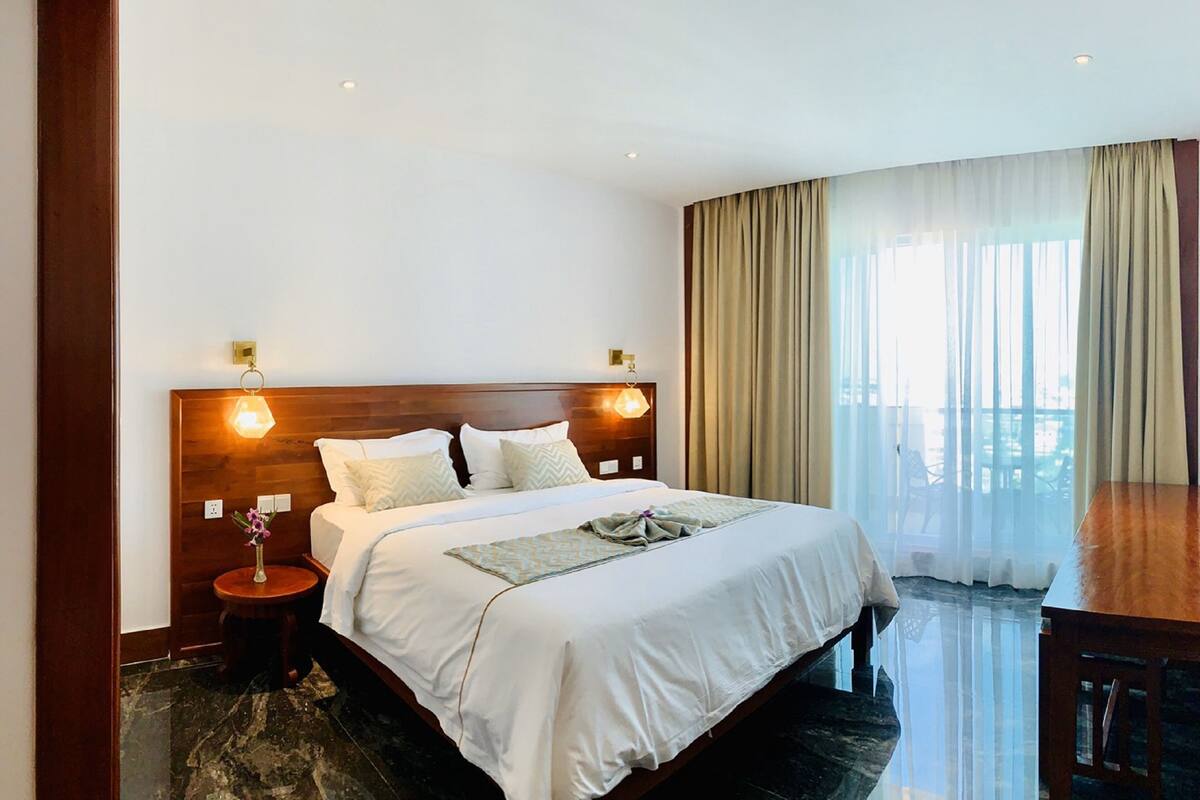 A hotel room with a large bed, wooden headboard, two bedside lamps, a small table with a vase, and a view of the outdoors through sheer curtains.