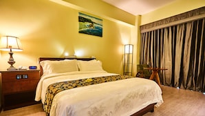 A hotel room with a bed, bedside table, lamp, painting, and curtains. - Serenti Hotel (Saipan)