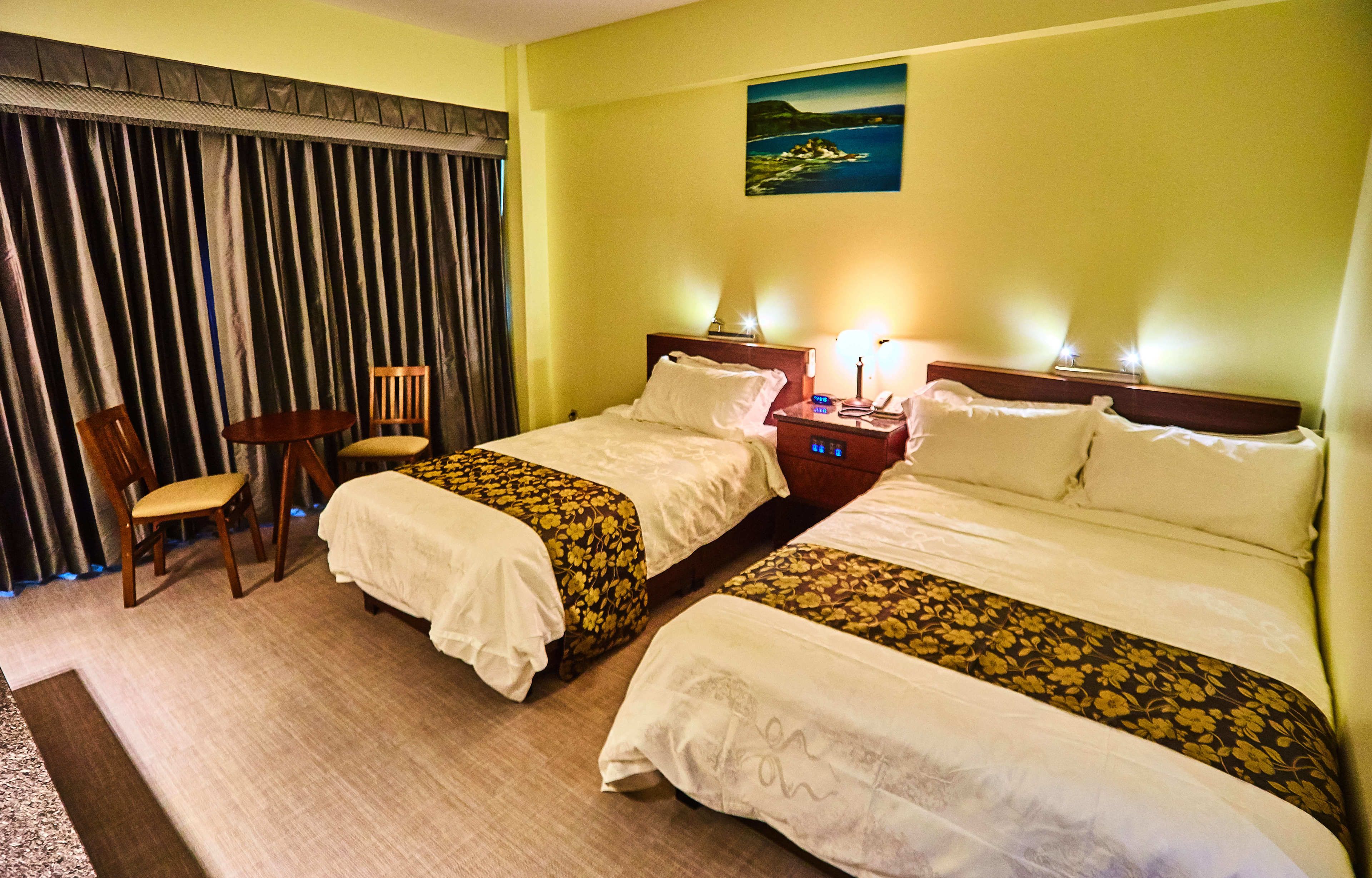 Habitación de hotel con dos camas, una silla, una mesita de noche con lámpara y un cuadro en la pared.